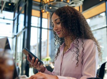 Woman sitting at restaurant table and scrolling on a mobile phone, with a laptop open on the table