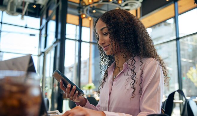 Woman sitting at restaurant table and scrolling on a mobile phone, with a laptop open on the table