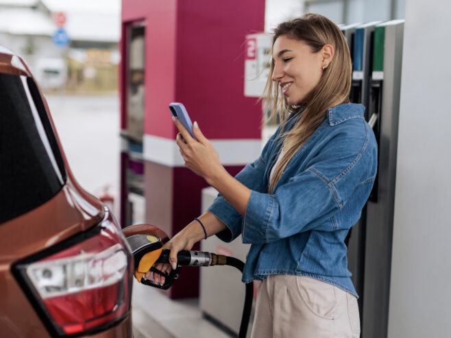 woman at gas station pumping gas while checking her phone